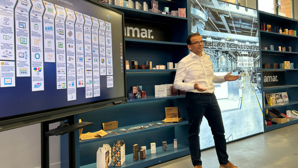 Un hombre de pie frente a una pantalla que muestra un diagrama de flujo, explicando conceptos relacionados con la impresión, en un ambiente de oficina moderno con estanterías llenas de productos.