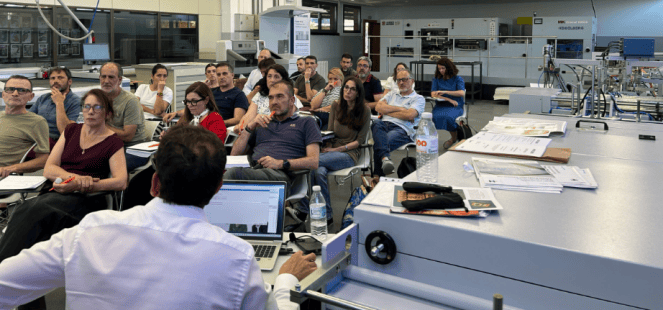 Una sala de clase con un grupo de personas sentadas, prestando atención a un presentador que se encuentra frente a una computadora portátil, en un entorno educativo o de capacitación.