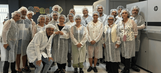 Grupo de personas posando dentro de una planta de producción, vistiendo batas y cofia, con un fondo que muestra maquinaria y el logo de '2Pack'.